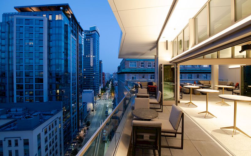 A modern urban balcony view at dusk with tables, chairs, and a glass railing overlooking tall buildings and a lit city street.