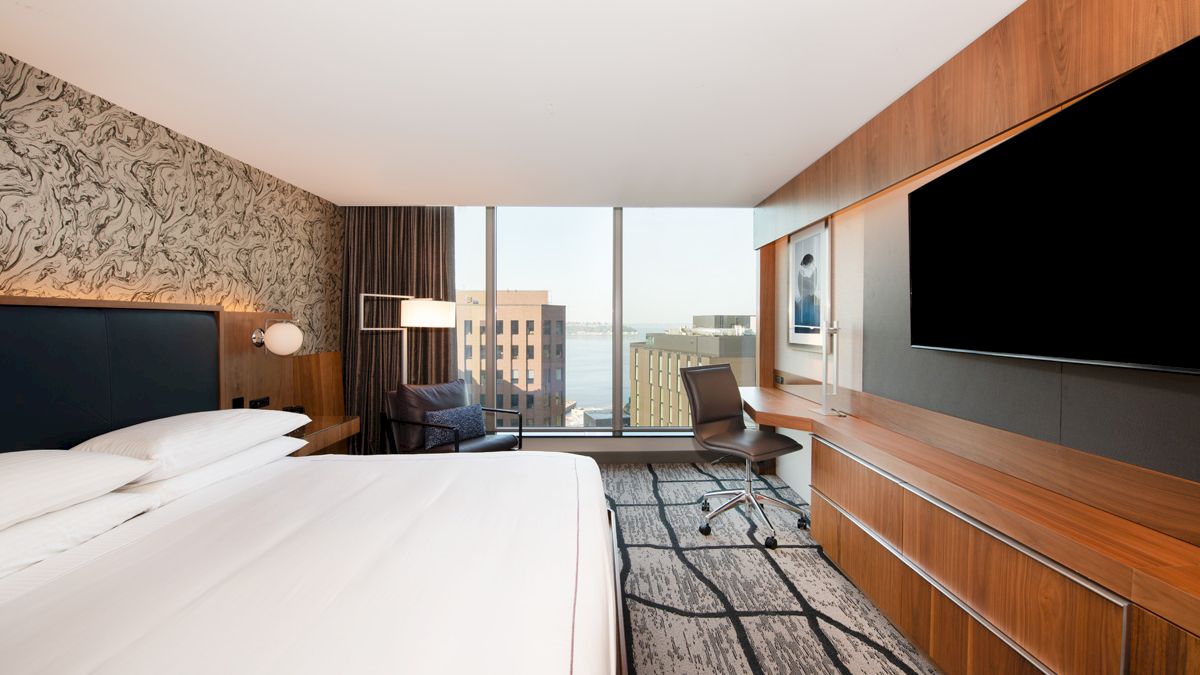 A modern hotel room with a large bed, wall-mounted TV, desk, chair, and a view of buildings and water through floor-to-ceiling windows.
