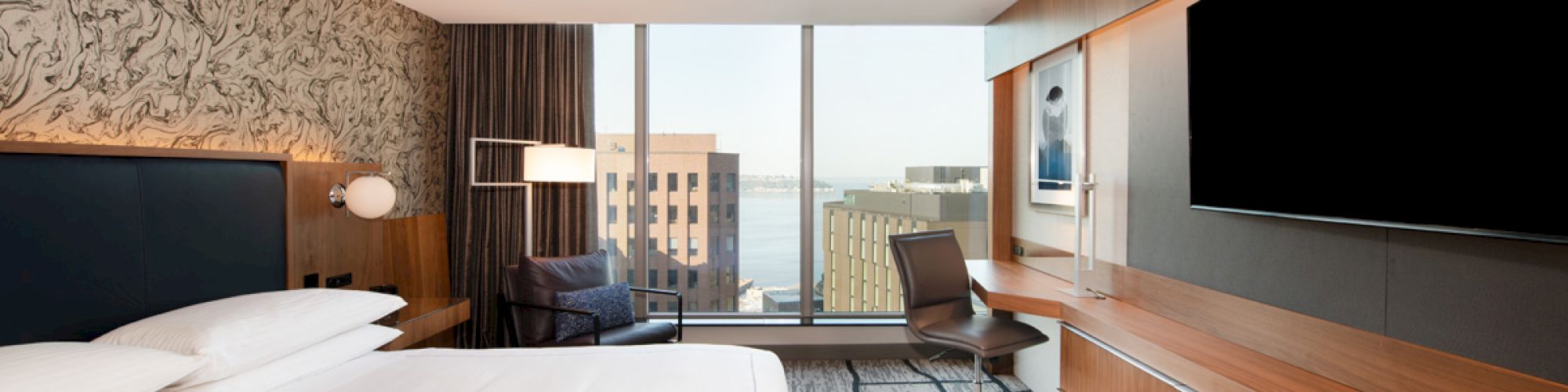 A modern hotel room with a large bed, wall-mounted TV, desk, chair, and a view of buildings and water through floor-to-ceiling windows.