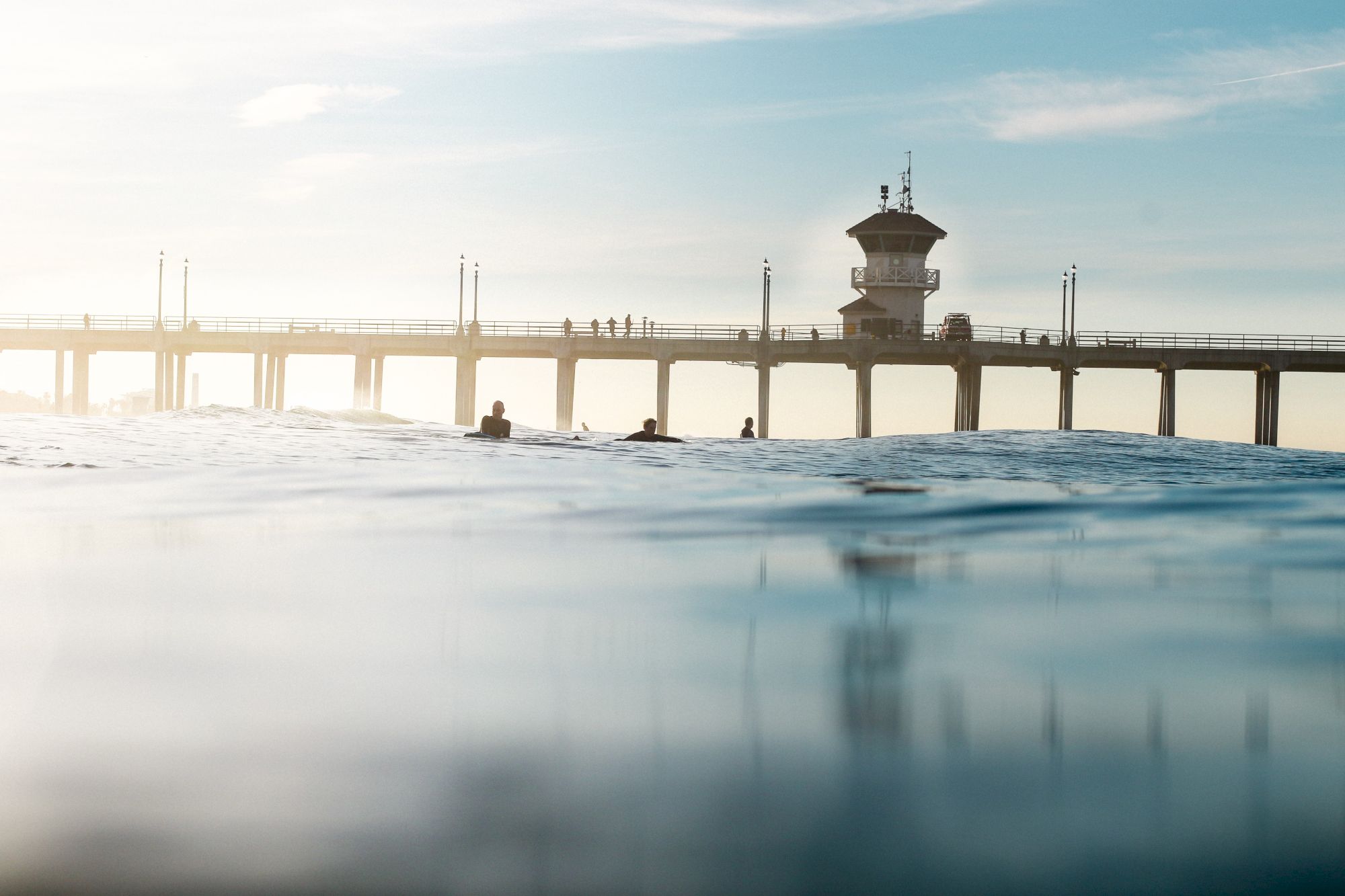 A tranquil view of a pier extending into the ocean with a group of surfers in the water, under a clear sky.